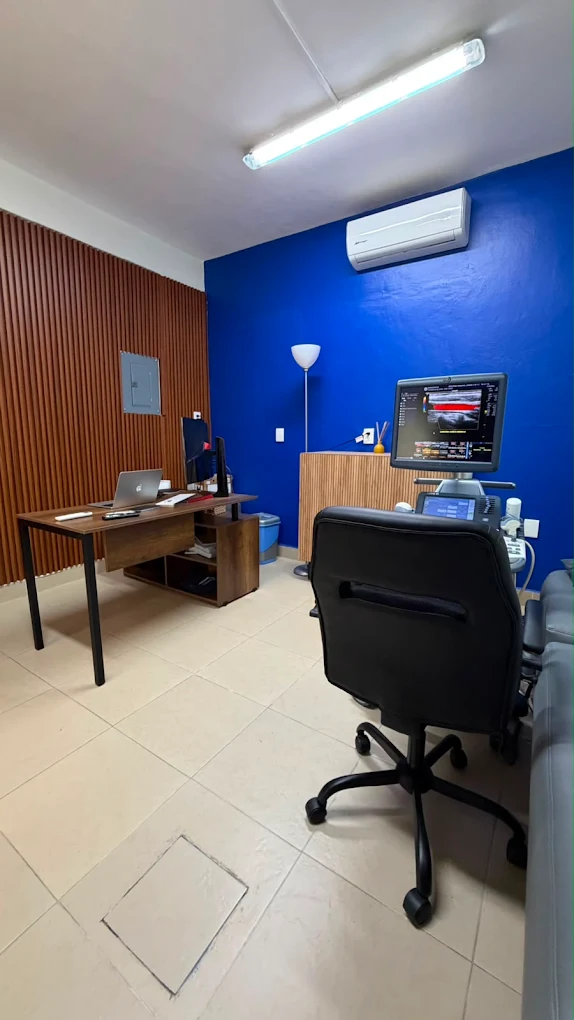 Consultorio de consulta: escritorio de madera, pared azul y equipo de ultrasonido con monitor